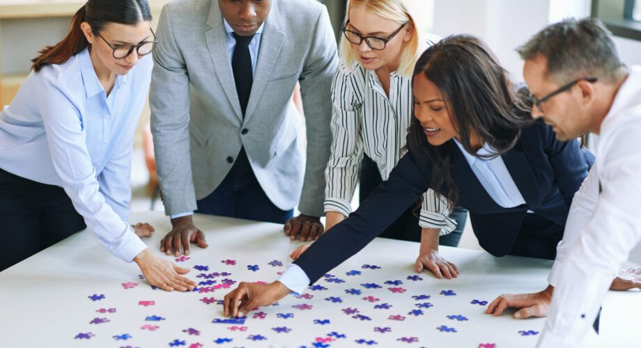 Diverse group of smiling businesspeople working together around a boardroom table in an office trying to solve a jigsaw puzzle