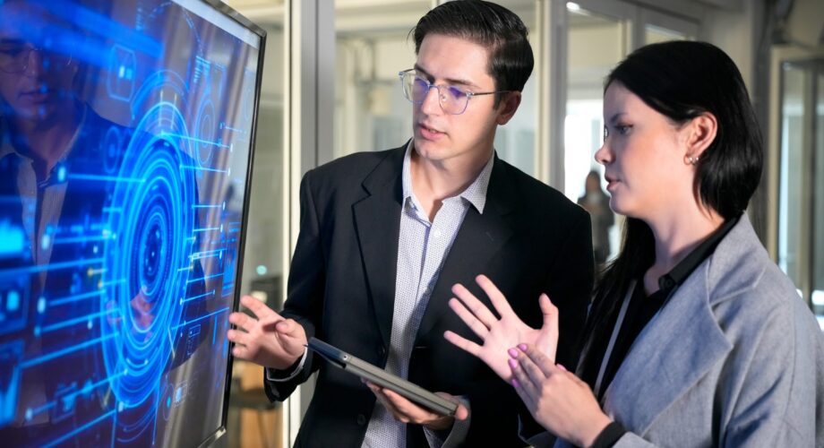 Two professionals are engaged in a conversation in an office. One person points at a screen while the other listens and gestures with hands. The atmosphere is focused on technology.