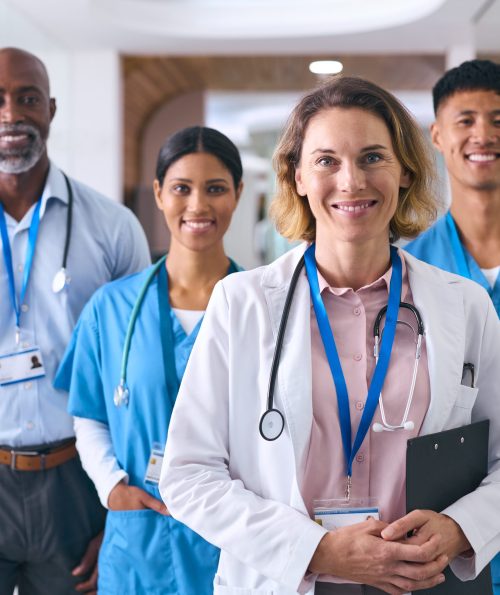 Portrait Of Smiling Multi Cultural Medical Team Wearing Scrubs And White Coats In Modern Hospital