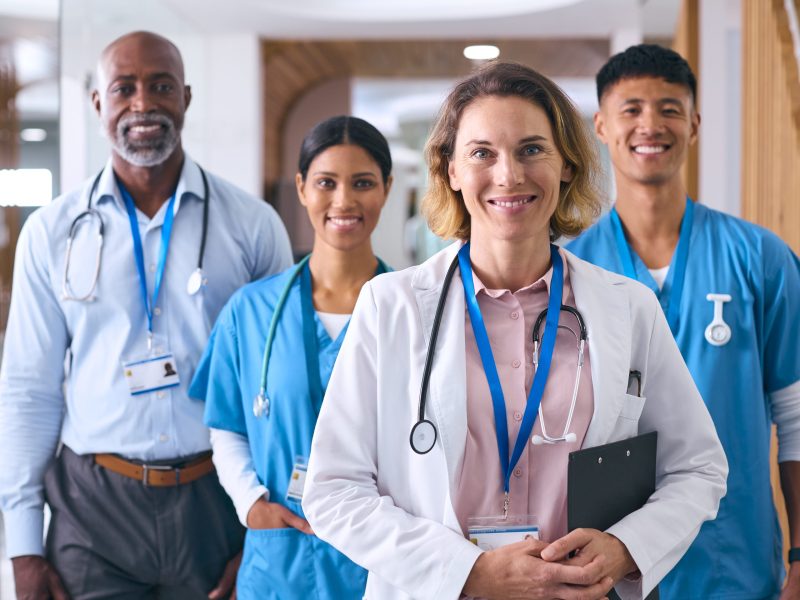Portrait Of Smiling Multi Cultural Medical Team Wearing Scrubs And White Coats In Modern Hospital