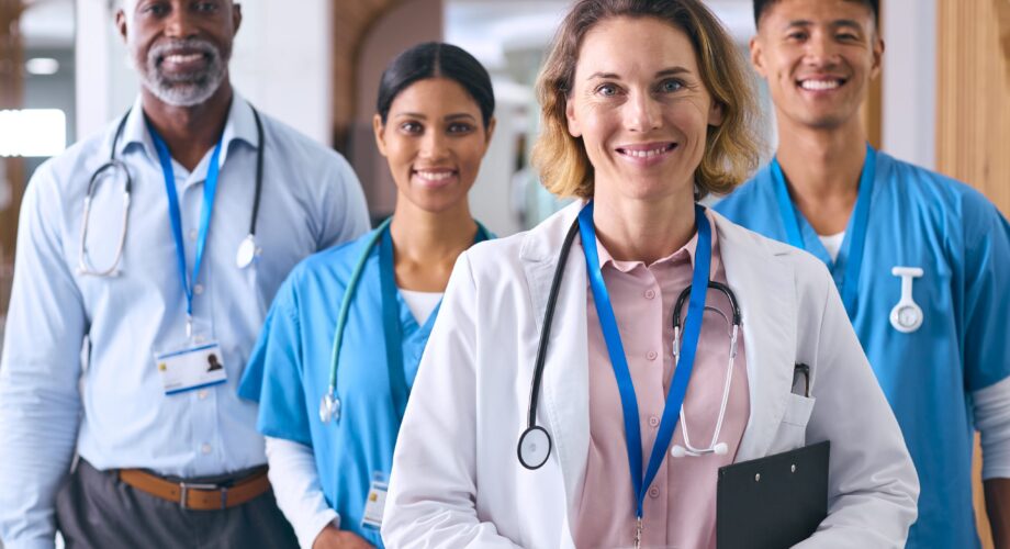 Portrait Of Smiling Multi Cultural Medical Team Wearing Scrubs And White Coats In Modern Hospital