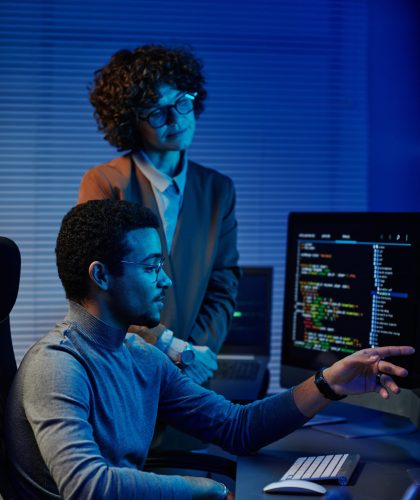 Confident IT engineer or software developer pointing at codes on computer screen while making presentation to female colleague