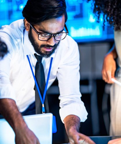 Diverse AI technology team including South Asian male and Black female professionals presents data on a large screen in a dark conference room.